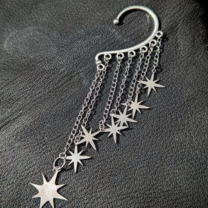May include: Silver ear cuff featuring a curved top that hooks over the ear. Multiple chains dangle from the cuff, each adorned with a star-shaped charm. The ear cuff is designed to be worn on the upper ear.