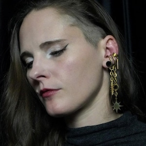 May include: A close-up of a person wearing a gold-colored ear gauge and dangling earrings. The earrings feature a starburst, chain, and circular design. The person is wearing a dark gray turtleneck.