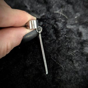 May include: A silver-colored ear cuff and a dangling bar earring. The ear cuff is a simple, rounded band. The earring features a small loop connecting to a long, straight bar. The jewelry is displayed against a dark, textured background.