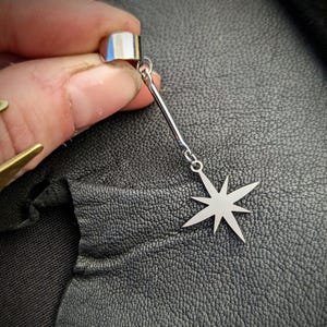 May include: A silver ear cuff with a dangling starburst charm. The ear cuff is a simple, polished metal band that wraps around the ear. The starburst charm is a flat, seven-pointed star.