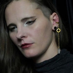 May include: A gold-colored ear cuff with a dangling chain and a clear crystal pendant. The ear cuff is attached to a black stud earring. The chain has a gold-colored ring in the middle.