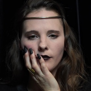 May include: A woman with dark lipstick and matte black nail polish. She wears a gold headband and a gold ring. Her hand is near her face. The background is black.