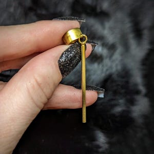 May include: A gold-colored earring with a cylindrical top and a long, straight, dangling bar. The earring is held between fingers with black glitter nail polish.