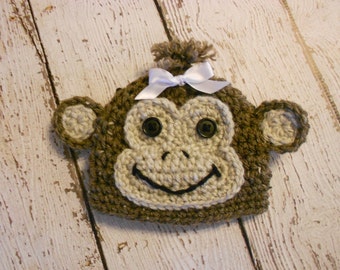 May include: A brown crocheted monkey hat with a white bow and black button eyes.