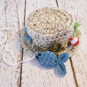 Puede incluir: Un sombrero de pesca tejido a crochet en tonos marrones y beige. El sombrero tiene un peque&ntilde;o pez azul sujeto a la parte delantera y un flotador rojo y blanco en el lateral.