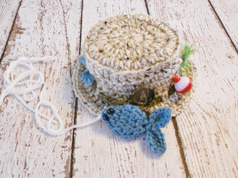 Puede incluir: Un sombrero de pesca tejido a crochet en tonos marrones y beige. El sombrero tiene un peque&ntilde;o pez azul sujeto a la parte delantera y un flotador rojo y blanco en el lateral.