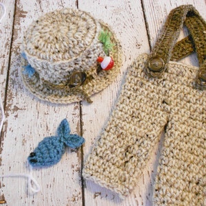 Puede incluir: Un conjunto de pesca de ganchillo para beb&eacute;, que incluye un gorro, un mono y un peque&ntilde;o pez azul. El gorro tiene un borde marr&oacute;n y una decoraci&oacute;n de se&ntilde;uelo de pesca verde y rojo. El mono tiene tirantes marrones y botones marrones. El pez est&aacute; sujeto a una cuerda blanca.
