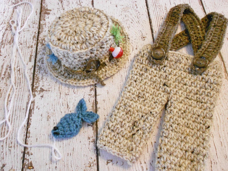 Puede incluir: Un conjunto de pesca de ganchillo para beb&eacute;, que incluye un gorro, un mono y un peque&ntilde;o pez azul. El gorro tiene un borde marr&oacute;n y una decoraci&oacute;n de se&ntilde;uelo de pesca verde y rojo. El mono tiene tirantes marrones y botones marrones. El pez est&aacute; sujeto a una cuerda blanca.