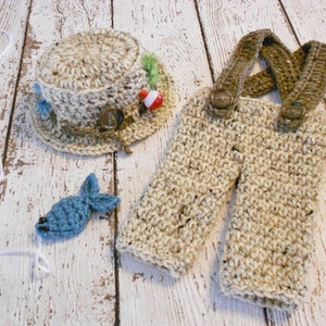 Puede incluir: Un conjunto de pesca de ganchillo para un beb&eacute;, que incluye un gorro, un mono y un peque&ntilde;o pez azul. El gorro tiene un flotador de pesca rojo y blanco y hojas verdes. El mono tiene tirantes marrones y dos botones. El conjunto est&aacute; colocado sobre una superficie de madera blanca.