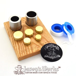 May include: A wooden base with two metal cylinders and five brass discs of varying sizes. The base has the text "Jason's Works" engraved on it. There is a black sticker with the text "Jason's Works" and a website address. Two rolls of tape, one blue and one white, are on the right side of the image.