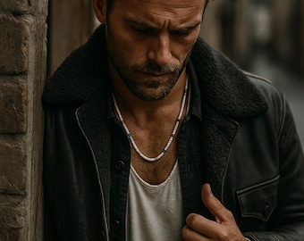 May include: A beaded necklace featuring small white cylindrical beads with occasional blue and brown accents, worn alongside a matching beaded bracelet on the wrist. The jewelry set is styled with a dark jacket, a dark button-down shirt, and a light undershirt.