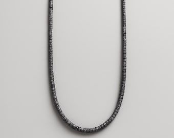 May include: A dark gray necklace composed of small, square beads. The beads are strung together to form a continuous chain. The necklace hangs against a neutral background, showcasing its simple, yet elegant design. This jewelry piece is suitable for everyday wear.