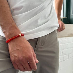 May include: A red carnelian beaded bracelet with a gold bead accent. The bracelet is worn on a person's wrist.
