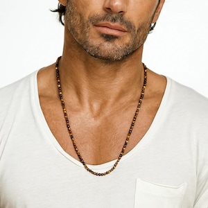 Può includere: Una collana di perline marroni e dorate. Le piccole perline rotonde sono infilate per formare una lunga catena. La persona indossa una maglietta bianca con scollo a V.