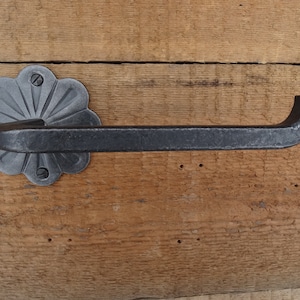 May include: Black wrought iron toilet paper holder with a decorative flower-shaped base. The holder is mounted on a wooden wall.