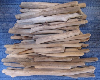 May include: A pile of weathered driftwood pieces in various sizes and shapes. The wood has a natural, light brown color with some pieces showing a silvery-gray hue. The arrangement is on a blue woven surface.