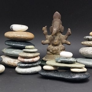 May include: A collection of balanced stone stacks in various sizes and colors, with a small bronze statue of a deity in the center. The stones range from light gray to dark gray and brown.