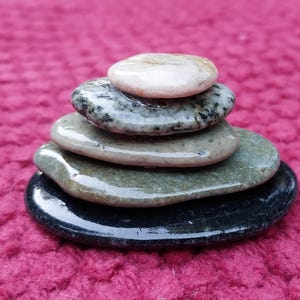 May include: A stack of five smooth, flat stones in varying shades of gray, green, and black. The top stone is a light beige color. The stones are arranged in a balanced tower, creating a natural, minimalist aesthetic.