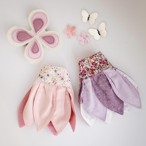 May include: A set of fabric accessories including a large butterfly-shaped piece with pink and cream layers, two small white butterflies, two small pink flowers, and two petal-shaped skirts with floral waistbands in shades of pink and purple.