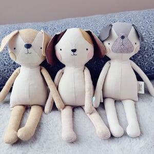 Peut inclure: Trois poupées en forme de chien en peluche, avec des corps beiges et des oreilles et traits du visage de couleurs différentes. Les chiens ont des détails faciaux cousus, dont des yeux et des nez noirs, et sont posés sur une surface douce et claire. Ces poupées sont un ajout charmant à toute chambre d'enfant.