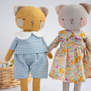 May include: Two handmade cat dolls. One is mustard yellow, wearing a blue gingham outfit with a white collar. The other is grey and wears a floral dress with a ruffled collar. Both dolls have pink cheeks and cat ears.
