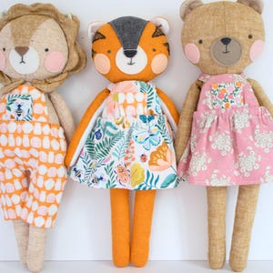 May include: Three fabric dolls: a lion, a tiger, and a bear. The lion wears orange overalls with a white pattern. The tiger has an orange body and a floral dress. The bear wears a pink floral dress. All dolls have rosy cheeks.