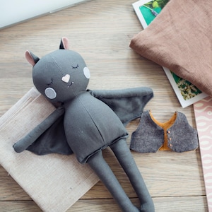 May include: A handmade gray bat doll with outstretched wings, a heart-shaped nose, and closed eyes, resting on a beige cloth. A small gray vest with an orange lining and a pink and white patterned notebook are nearby.