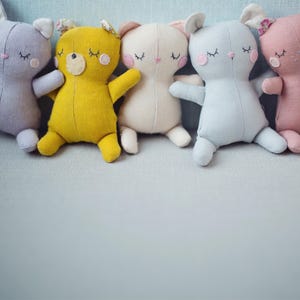 May include: Five handmade plush animal toys in various colors: lavender, yellow, cream, light blue, and pink. Each toy has closed eyes, small arms, and ears. The toys are arranged in a row on a light blue surface.