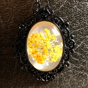 Puede incluir: Un collar con un colgante de metal negro y un camafeo ovalado de resina transparente con flores amarillas prensadas.