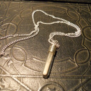 May include: A silver chain necklace with a small glass vial pendant filled with white sand. The necklace is lying on a brown leather surface with an intricate pattern.