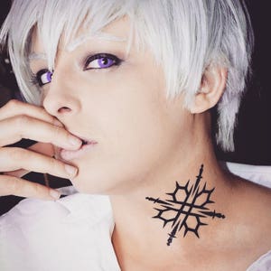 May include: A person with white hair and purple eye contacts is wearing a white shirt. They have a black tattoo of a geometric design on their neck.