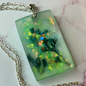 May include: A rectangular pendant necklace with a silver chain. The pendant is a light green resin, encasing dried flowers and iridescent glitter. The chain is a delicate silver link style, suitable for everyday wear.