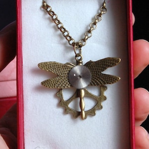 May include: A gold-toned dragonfly pendant necklace with a gear design and a small, round, silver-toned clock face. The necklace is displayed in a red box with white lining and the text "NatiniNatini" printed on the bottom.