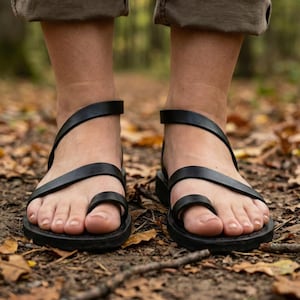 May include: Black leather sandals with a toe loop and ankle strap. The sandals have a flat sole and are worn outdoors. The image shows the sandals on a person's feet, with a blurred background of leaves and trees.