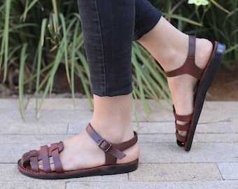 Peut inclure: Sandales en cuir marron avec un design de lanières tissées et une boucle réglable. Les sandales sont portées par une personne.