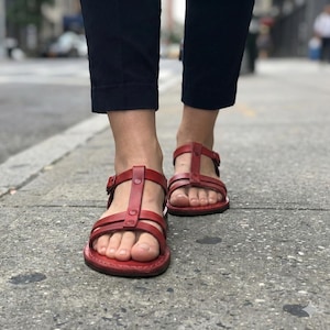 May include: Red leather sandals with multiple straps and buckles. The sandals have a flat sole and are being worn on a city sidewalk. The sandals are a vibrant red color, and the image is taken from a low angle.