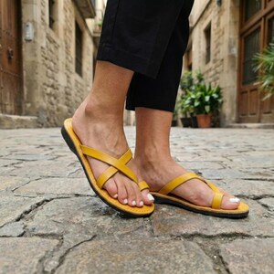 May include: Yellow leather sandals with crisscross straps. The sandals have a black sole and are worn on a cobblestone street. The sandals are a bright, summery color and appear to be comfortable.