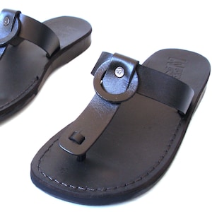 Puede incluir: Un par de sandalias de cuero negro con una hebilla de anillo y un diseño simple. Las sandalias tienen una suela plana y están hechas de cuero de alta calidad.
