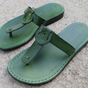 May include: A pair of green leather sandals with a buckle strap and a stitched edge. The sandals have a simple design and are made of high-quality leather.