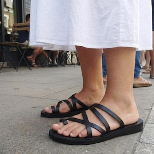May include: Black leather sandals with multiple straps and a toe loop. The sandals have a flat sole. The person is wearing a white dress. The sandals are being worn outdoors.