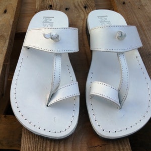 K&ouml;nnte beinhalten: Ein Paar wei&szlig;e Ledersandalen mit schlichtem Design. Die Sandalen haben einen Riemen, der zwischen den Zehen verl&auml;uft, und einen Riemen, der um den Kn&ouml;chel geht. Die Sandalen sind aus weichem Leder gefertigt und bieten einen bequemen Sitz.
