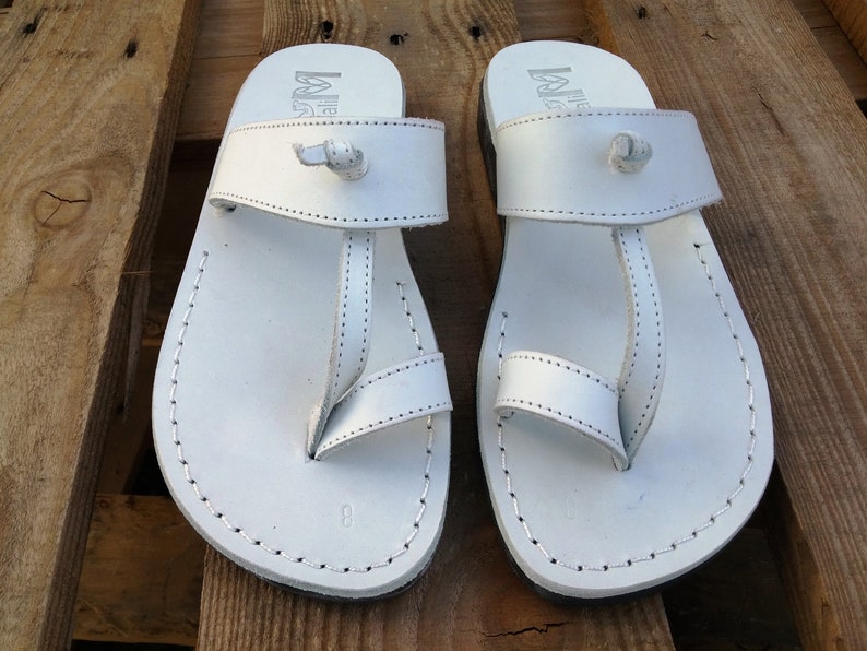 K&ouml;nnte beinhalten: Ein Paar wei&szlig;e Ledersandalen mit schlichtem Design. Die Sandalen haben einen Riemen, der zwischen den Zehen verl&auml;uft, und einen Riemen, der um den Kn&ouml;chel geht. Die Sandalen sind aus weichem Leder gefertigt und bieten einen bequemen Sitz.