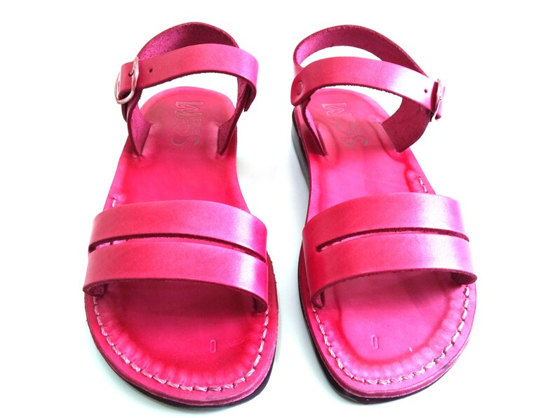 May include: A pair of pink leather sandals with an ankle strap and buckle fastening. The sandals have a simple design with a single strap across the toe.
