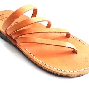 May include: Tan leather sandals with multiple straps and a white stitched sole. The sandals have a simple design and are made of high-quality leather.