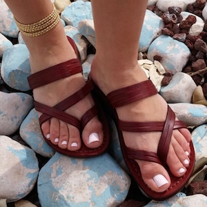 Op de afbeelding: Een paar bruine leren sandalen met gekruiste banden. De sandalen worden gedragen op de voeten van een persoon, die op een bed van stenen staat.