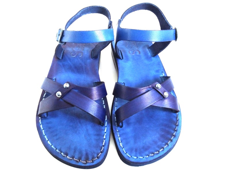 May include: A pair of blue leather sandals with a cross-strap design. The sandals have adjustable ankle straps and are stitched with white thread. The leather has a gradient effect, transitioning from a lighter to a darker shade of blue.