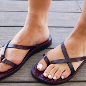 May include: Brown leather sandals with criss-cross straps and a thong toe. The sandals have a flat sole and are worn on a person's feet.
