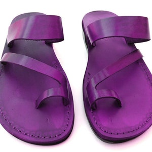 May include: A pair of purple leather sandals with a toe loop and two straps. The sandals have a stitched edge and a flat sole.