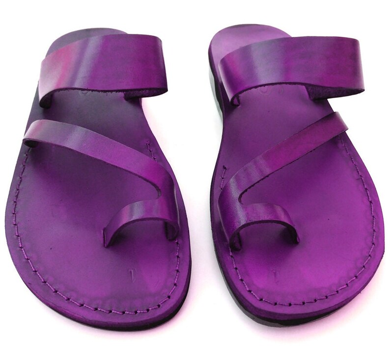 May include: A pair of purple leather sandals with a toe loop and two straps. The sandals have a stitched edge and a flat sole.