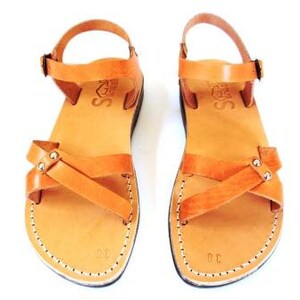 May include: A pair of tan leather sandals with a cross-strap design and ankle straps. The sandals have a stitched sole and a buckle closure. The sandals are size 38. The sandals are displayed on a white background.